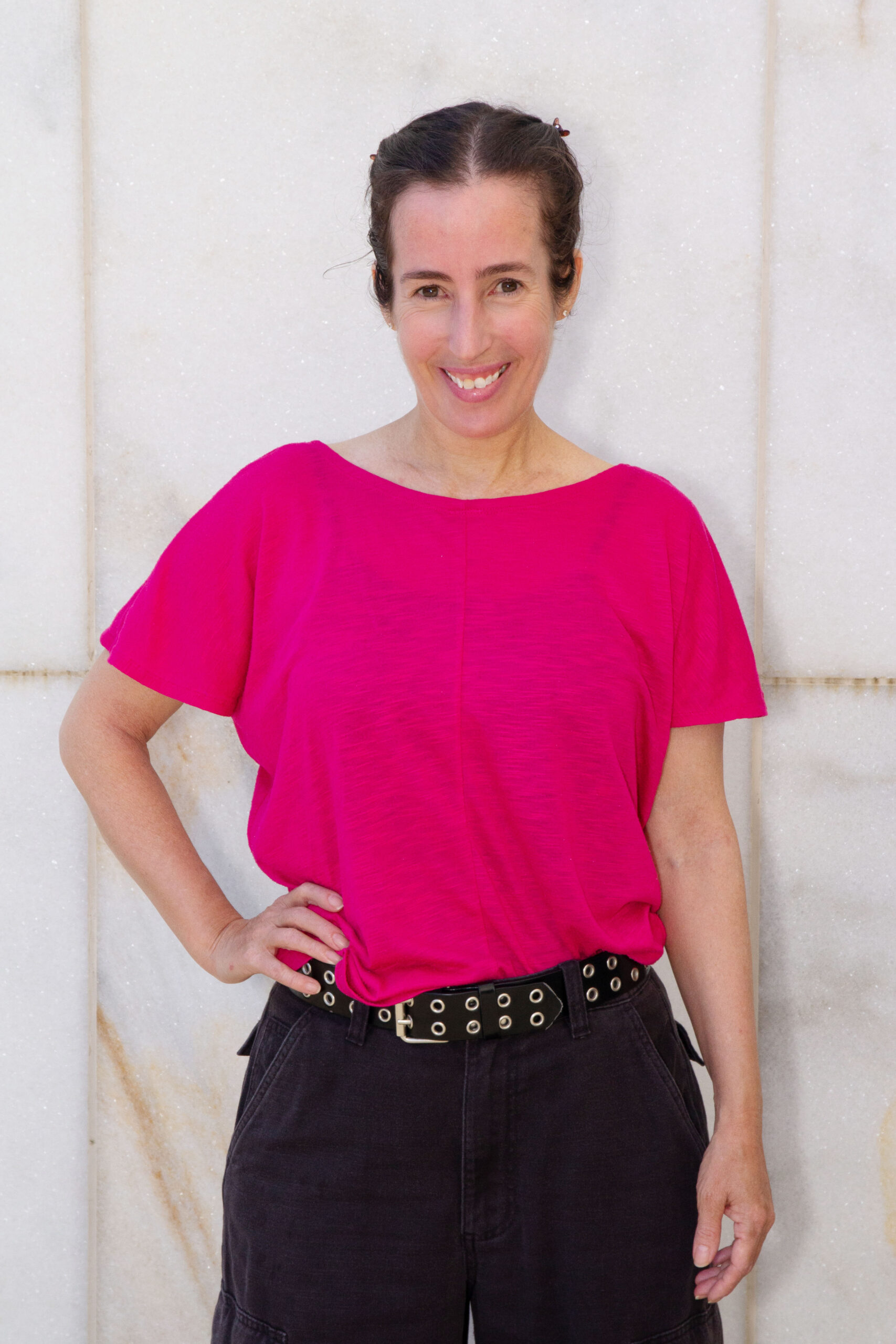 A full colour image of Felicity Hamill wearing a bright pink top and dark trousers, standing in front of a pale marbled wall, smiling directly into the lens.
